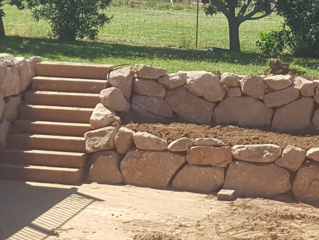 Rock Wall 1 rock wall Rustic stone staircase nestled between sections of a retaining rock wall, surrounded by lush greenery and trees in a natural landscape.